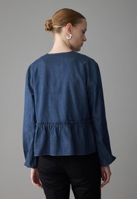 Blouse en denim foncé avec une taille cintrée, des manches longues et un col arrondi. Texture lisse avec un détail froncé à l'arrière.