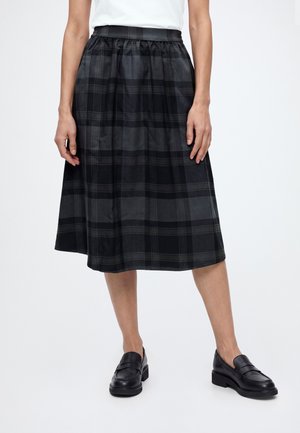 Mulher vestindo uma saia midi xadrez preta e cinza com cós elástico e mocassins pretos, em pé contra um fundo branco simples.