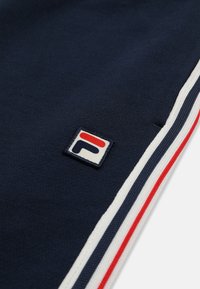 Tissu bleu marine avec bordure rayée blanche, marine et rouge, et un petit patch logo carré présentant des formes stylisées rouges et marine.