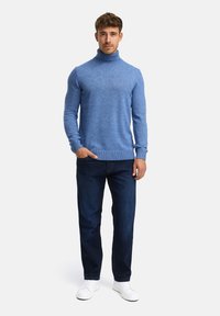 Hellblauer Rollkragenpulli aus strukturiertem Material, kombiniert mit dunkelblauen Jeans und weißen Sneakers, das Model steht in einer neutralen Pose.