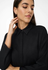 Zwarte langslavige shirt met een knoopsluiting bij de kraag, gemaakt van zachte stof en voorzien van een plooi aan de voorkant. Model toont eenvoudige sieraden en een ontspannen houding.