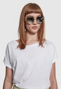 Ochelari de soare cu o formă geometrică de tip cat-eye, lentile reflectorizante, ramă din metal ușor și detalii subtile pe sprânceană. Fundal de tricou alb.