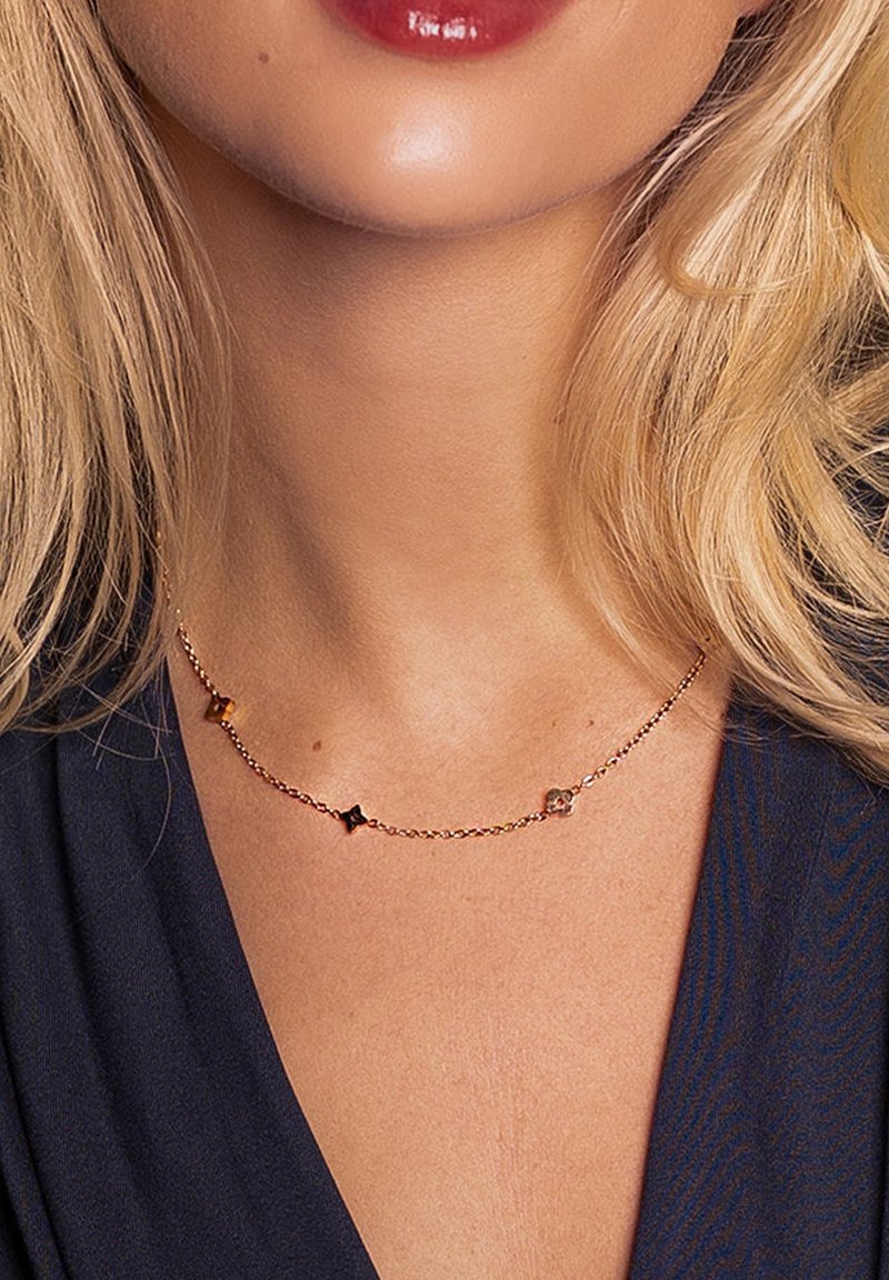 Collier délicat en chaîne d'or avec trois petits pendentifs en forme de fleurs, porté autour du cou d'une femme aux cheveux blonds et vêtue de vêtements sombres.
