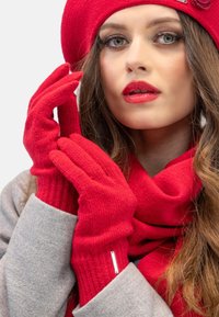 Gants en tricot rouge avec texture côtelée, associés à une écharpe rouge assortie et un manteau gris. Présentent un accent argenté sur les gants.