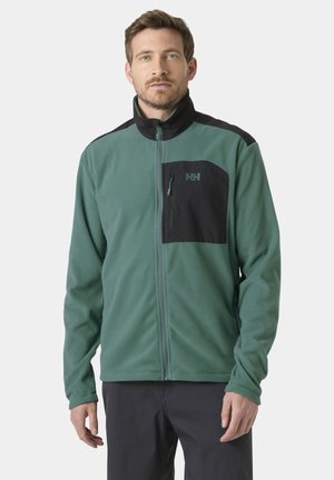 Hombre que lleva una chaqueta de forro polar verde con cremallera, con bolsillo negro en el pecho y paneles en los hombros, de pie frente a un fondo liso.