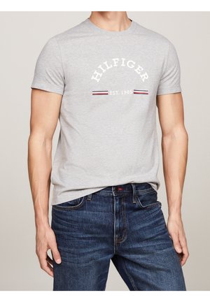 Męski tors w jasnoszarej koszulce z krótkim rękawem z napisem „Hilfiger EST. 1985” i paskowanymi akcentami, w połączeniu z ciemnoniebieskimi dżinsami.