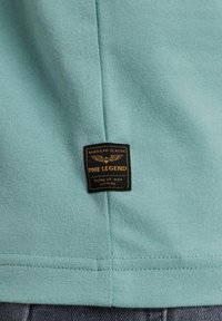 Majico zeleni bombažni T-shirt z črno in zlato oznako blagovne znamke. Tkanina je gladka, šivi pa so urejeni ob robu in na strani.