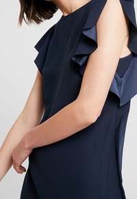 Vrouw in een mouwloze marineblauwe jurk met gerimpelde schouderdetails, met een licht gebogen arm over het lichaam tegen een eenvoudige achtergrond.