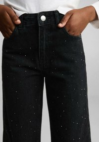 Černé džíny z denimu s vysokým pasem, velkými předními kapsami a malými stříbrnými detaily rozptýlenými po látce. Stříbrný knoflík v pase.