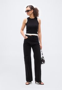 Frau trägt ein schwarzes ärmelloses Oberteil, hoch taillierte, weit geschnittene schwarze Hose, schwarze Sandalen, Sonnenbrille mit Sternakzenten, hält eine schwarz-weiß gemusterte Handtasche.