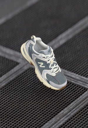 Baskets de sport grises en matériaux mesh et synthétiques, avec des lacets blancs et une semelle beige, présentées sur une surface texturée.