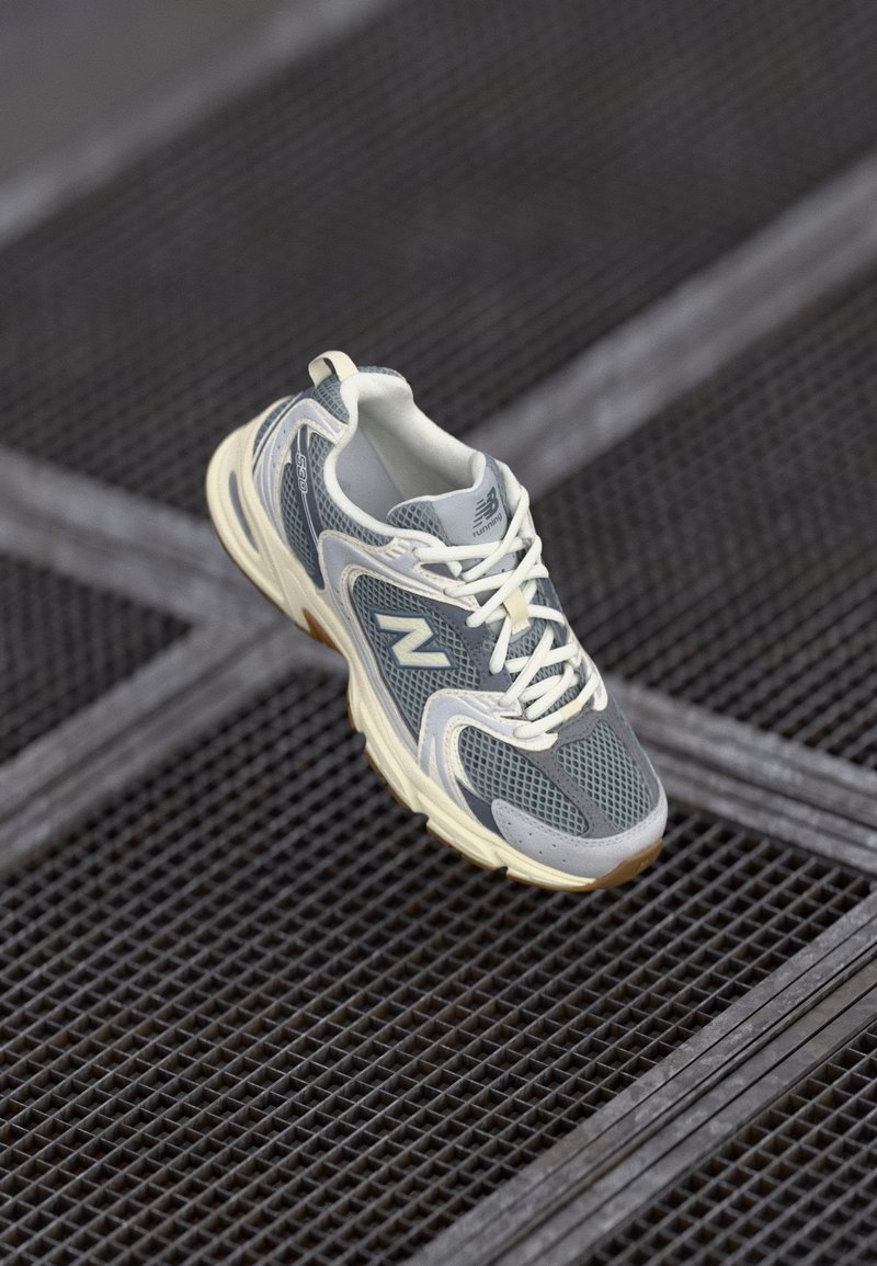 Gray athletic shoe with mesh and synthetic materials, featuring white laces and a beige sole, showcased on a textured surface.