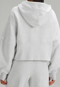 Sweat à capuche gris court avec capuche, manches larges et tissu texturé. Présente des coutures aux épaules et un détail logo à l'arrière près du col.