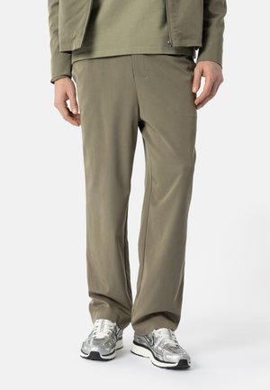 Mann iført olivengrønne bukser og matchende jakke med grønn skjorte, stående i sølv- og hvite joggesko på hvit bakgrunn.