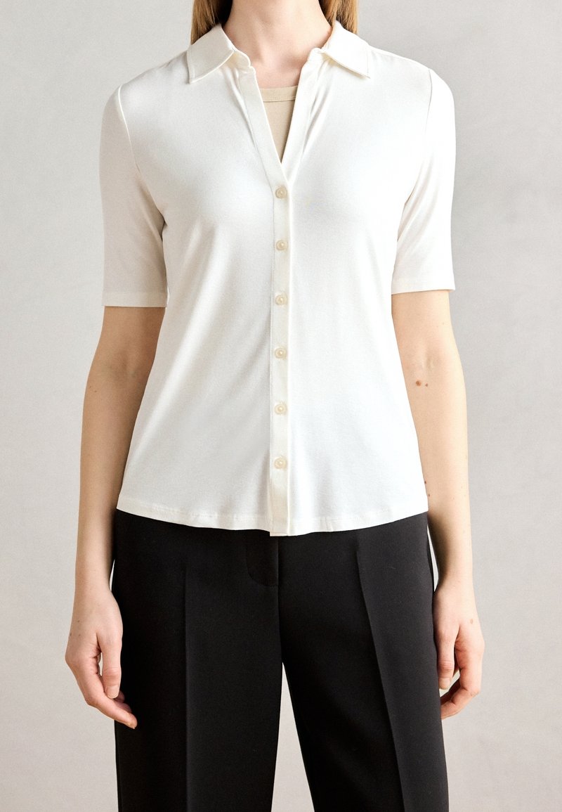 Femme portant une chemise blanche à manches courtes avec col et pantalon noir, montrée des épaules aux hanches sur un fond neutre.