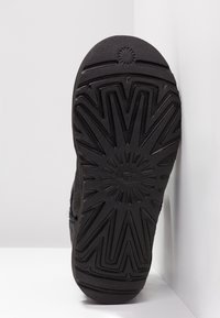 Semelle en caoutchouc noire d'une botte avec des motifs en zigzag et des lignes rayonnantes, placée debout contre un mur d'angle blanc.