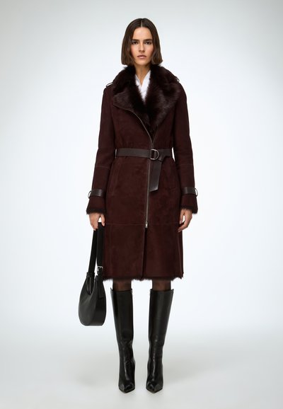 Horčicovočervený semišový kabát s umelým kožušinovým golierom, opaskom v páse, zipsovým zapínaním a čiernymi koženými detailmi. Kombinovaný s čiernymi čižmami po kolená.