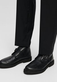Black leather shoes with a rounded toe, buckle accent, and rubber sole, paired with black pants, showcasing a polished design.