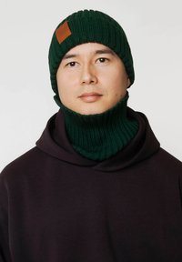 Zielona dzianinowa czapka i komin wykonane z prążkowanego materiału, z brązową skórzaną metką. Noszone na ciemnej bluzie z kapturem.