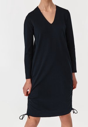 Frau trägt ein langärmliges schwarzes Kleid mit V-Ausschnitt und verstellbaren Kordeln am Saum, steht vor einem einfarbigen Hintergrund.