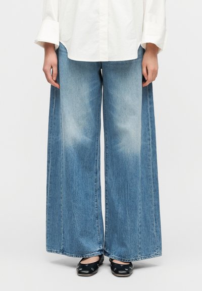 Παντελόνι από τζιν με φαρδιά ποδαρίνα σε ανοιχτό μπλε, με ξεθωριασμένη υφή. Συνδυασμένο με λευκό πουκάμισο με κουμπιά και μαύρα μποτάκια με επίπεδη σόλα.