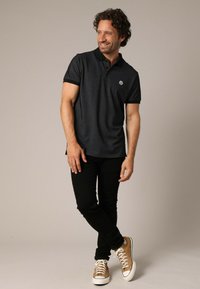 T-shirt polo noir avec un motif texturé, manches courtes et un petit logo sur la poitrine, associé à un jean noir et des baskets beiges.
