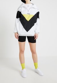 Biała bluza typu pulower z czarno-żółtym geometrycznym wzorem; do tego czarne szorty i białe trampki; żółte skarpetki.
