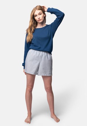 Blaues Langarmshirt und gestreifte Shorts. Das Shirt hat einen strukturierten Stoff; die Shorts haben eine lockere Passform mit einem elastischen Bund. Barfuß.