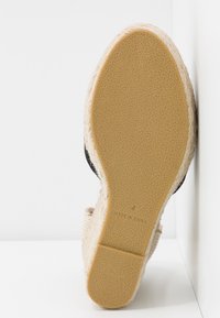 Semelle de chaussure de style espadrille avec un fond en caoutchouc texturé couleur tan et une section supérieure en jute tissé. Accents noirs visibles sur les côtés.