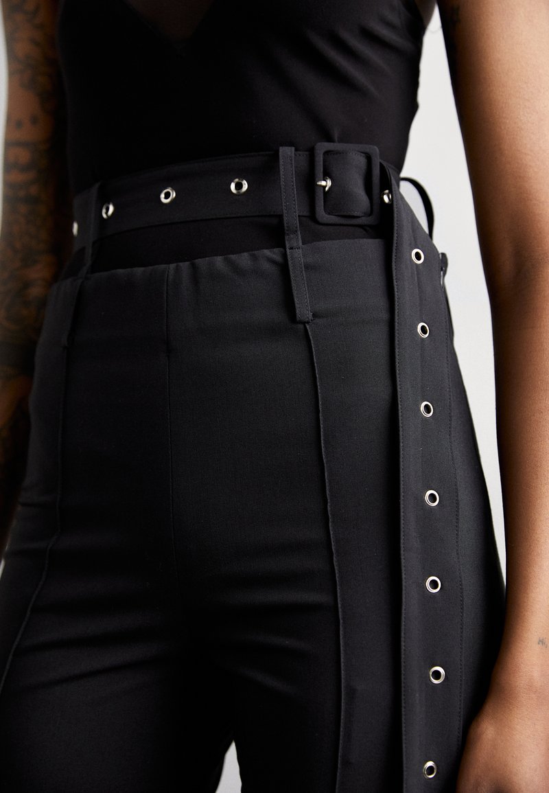 Pantalons noirs taille haute avec une large ceinture en métal et des détails de pression sur les côtés, fabriqués à partir d'un tissu lisse avec une coupe ajustée.