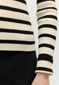 Gestreifter Ripp-Pullover in Creme und Schwarz mit langen Ärmeln; hat ein körperbetontes Design und einen Rippstrick, der an der Taille und am Handgelenk endet.