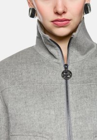 Graue Fleecejacke mit hohem Kragen, schwarzem Reißverschluss und rundem Metallakzent. Glatte Textur mit strukturiertem Design und Steppnähten.