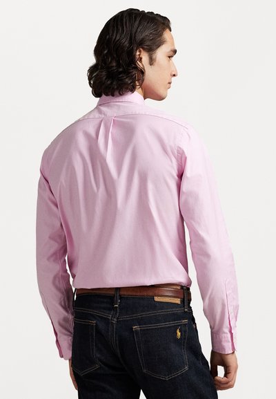 Camicia a maniche lunghe rosa chiaro in cotone, con colletto classico e piega sul retro, abbinata a jeans in denim scuro e una cintura marrone.