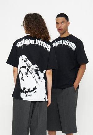 Δύο μοντέλα φορούν μαύρα T-shirts Maison Vienna με λευκό γοτθικό κείμενο· το ένα παρουσιάζει ένα μεγάλο λευκό γραφικό λύκου στην πλάτη, και τα δύο συνδυάζονται με γκρι φαρδιά παντελόνια.