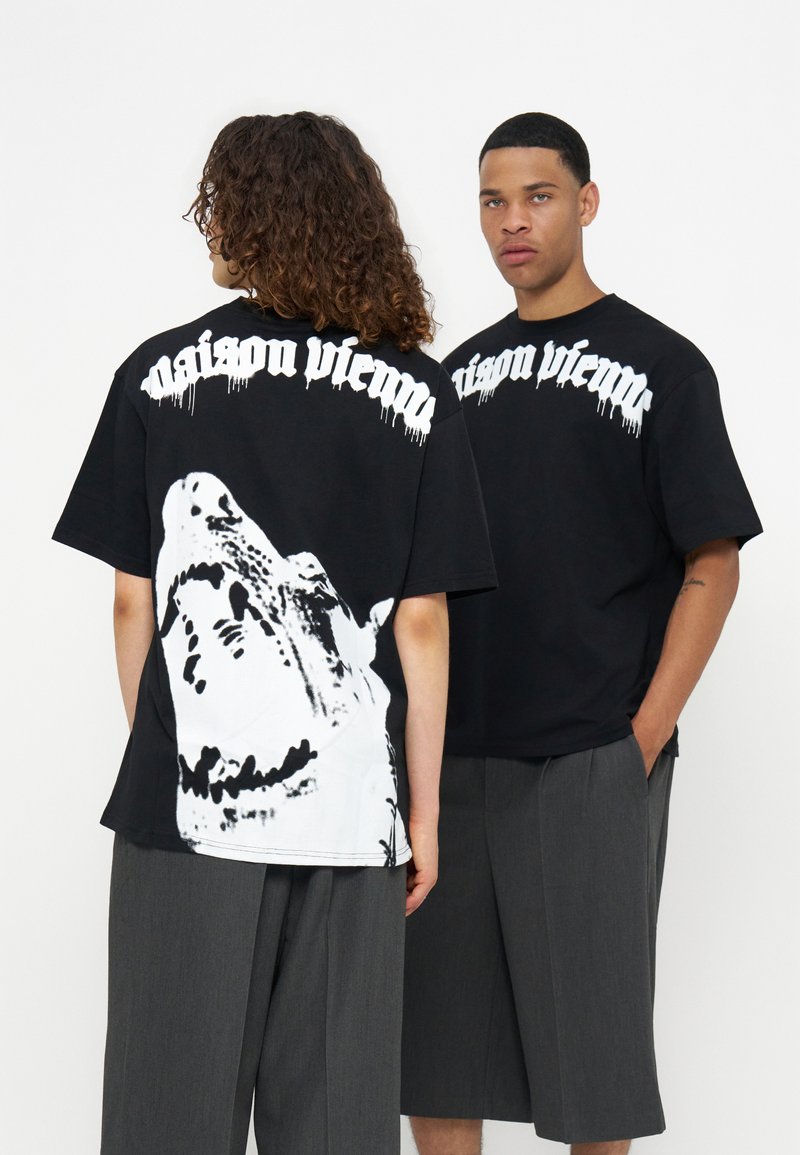 Δύο μοντέλα φορούν μαύρα T-shirts Maison Vienna με λευκό γοτθικό κείμενο· το ένα παρουσιάζει ένα μεγάλο λευκό γραφικό λύκου στην πλάτη, και τα δύο συνδυάζονται με γκρι φαρδιά παντελόνια.