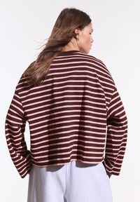 Femme aux longs cheveux bruns portant une chemise ample à rayures marron et rose à manches longues et un pantalon gris clair, vue de dos sur fond blanc.
