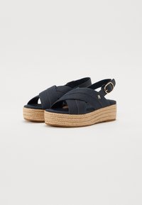 Sandalias de cuña de lona en color navy con diseño de tiras cruzadas, plataforma de cuerda de yute y correa ajustable para el tobillo con un acento de hebilla en tono dorado.