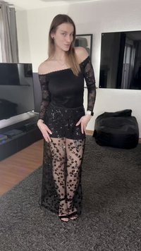 Black off-the-shoulder top, embellished sheer black skirt with floral sequin pattern, and strappy black sandals. Smooth fabric, elegant design.