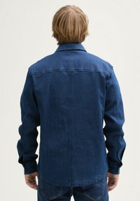 Jeansowa kurtka w ciemnoniebieskim kolorze, z kołnierzykiem, długimi rękawami i detalami na guzikach. Tkanina wygląda na teksturowaną i solidną.