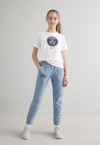 Jonge persoon staand met handen in zakken, draagt een wit Paris Saint-Germain T-shirt en blauwe joggingbroek met witte sneakers.