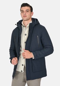 Giacca con cappuccio blu navy con chiusura a zip, due tasche sul petto e una grande tasca frontale. Presenta una texture trapuntata e polsini regolabili. Design leggero.