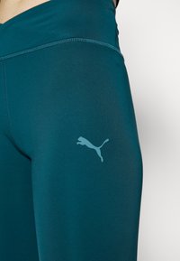 Turkusowe legginsy sportowe wykonane z gładkiego, elastycznego materiału. Posiadają małe, jasnoniebieskie logo Pumy na udzie oraz przeszywane szwy.