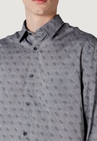 Camisa abotonada gris con una textura estampada de pequeñas formas de diamante y logotipos repetidos. Incluye un bolsillo en el pecho y botones negros.