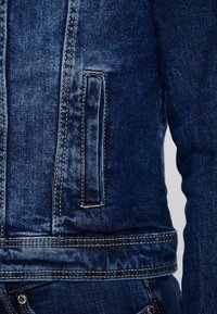 Tmavě modrá džínová bunda s texturovaným povrchem, viditelným prošitím a diagonální klopovou kapsou na levé straně.