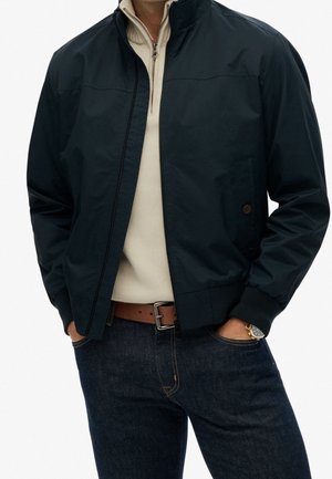 Homme portant une veste noire zippée sur un pull beige à fermeture éclair au quart, un jean foncé et une ceinture marron, les mains dans les poches.