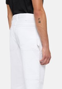 Hombre con pantalones blancos y camisa negra, mostrando el bolsillo trasero, el bolsillo lateral y un tatuaje en el antebrazo que dice "15:28".