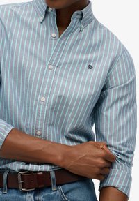Lichtblauw en mintgroen gestreept overhemd met knopen, een kraag en lange mouwen, en een klein logo. Draagt een bruine leren riem.