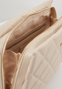 Borsa sintetica beige trapuntata con chiusura a zip dorata, caratterizzata da una texture liscia e un ampio interno foderato con una tasca aggiuntiva.