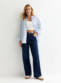 Lichtblauw en wit gestreepte overhemd met knopen, gedragen over een witte crop top, gecombineerd met hoge, donkere denim wijde broek en beige schoenen.