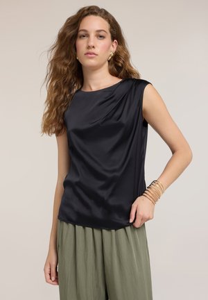Kvinde med bølget brunt hår iført sort satin top uden ærmer, olivengrøn plisseret nederdel, gyldne hoop-øreringe og stabler af gyldne armbånd.